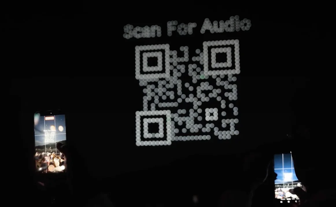 Drones forming a scannable Scan For Audio QR code over a night audience filming on their phones